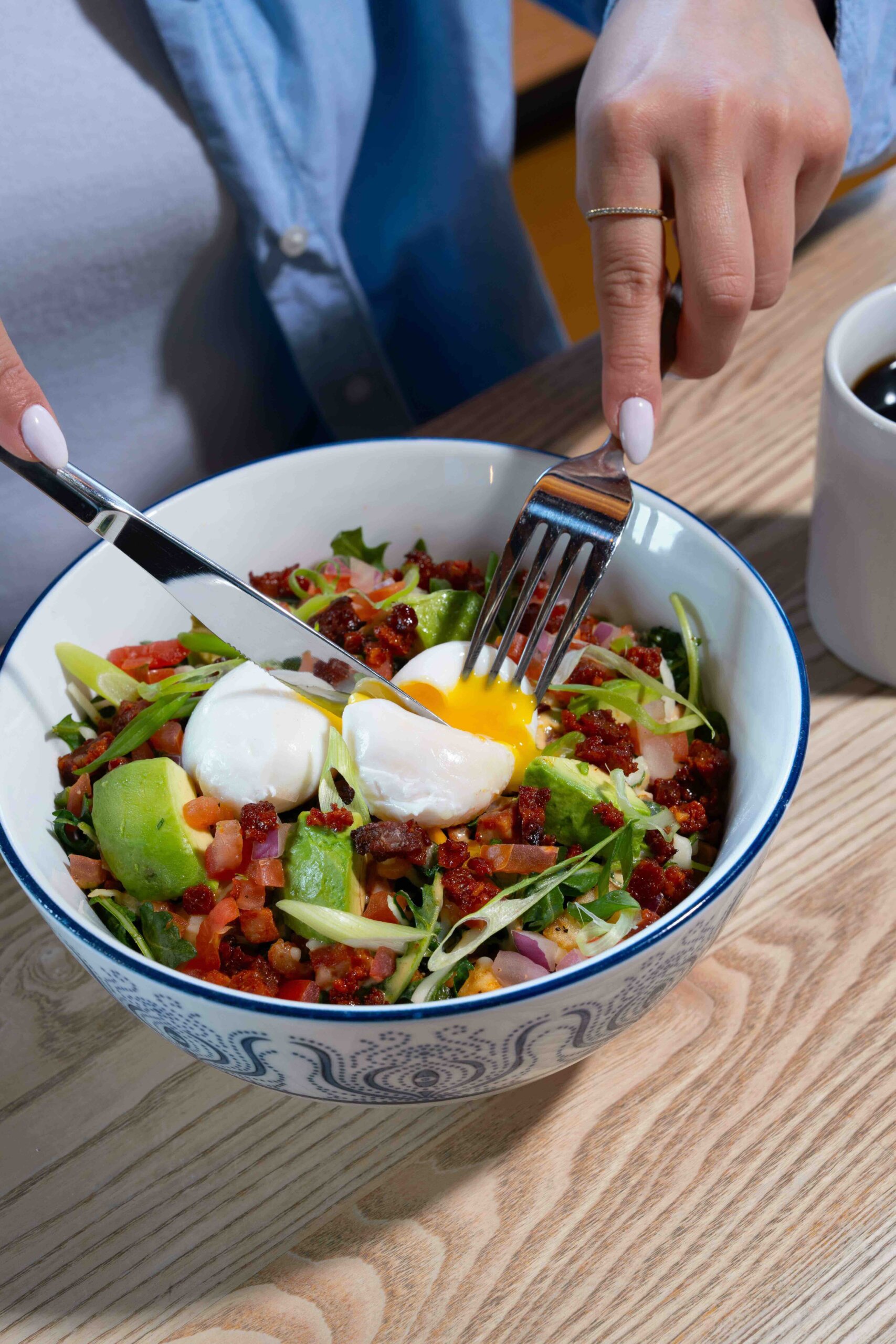 CRAFT Beer Market A person cuts into a poached egg atop a salad with avocado, diced vegetables, and greens in a patterned bowl on a wooden table.