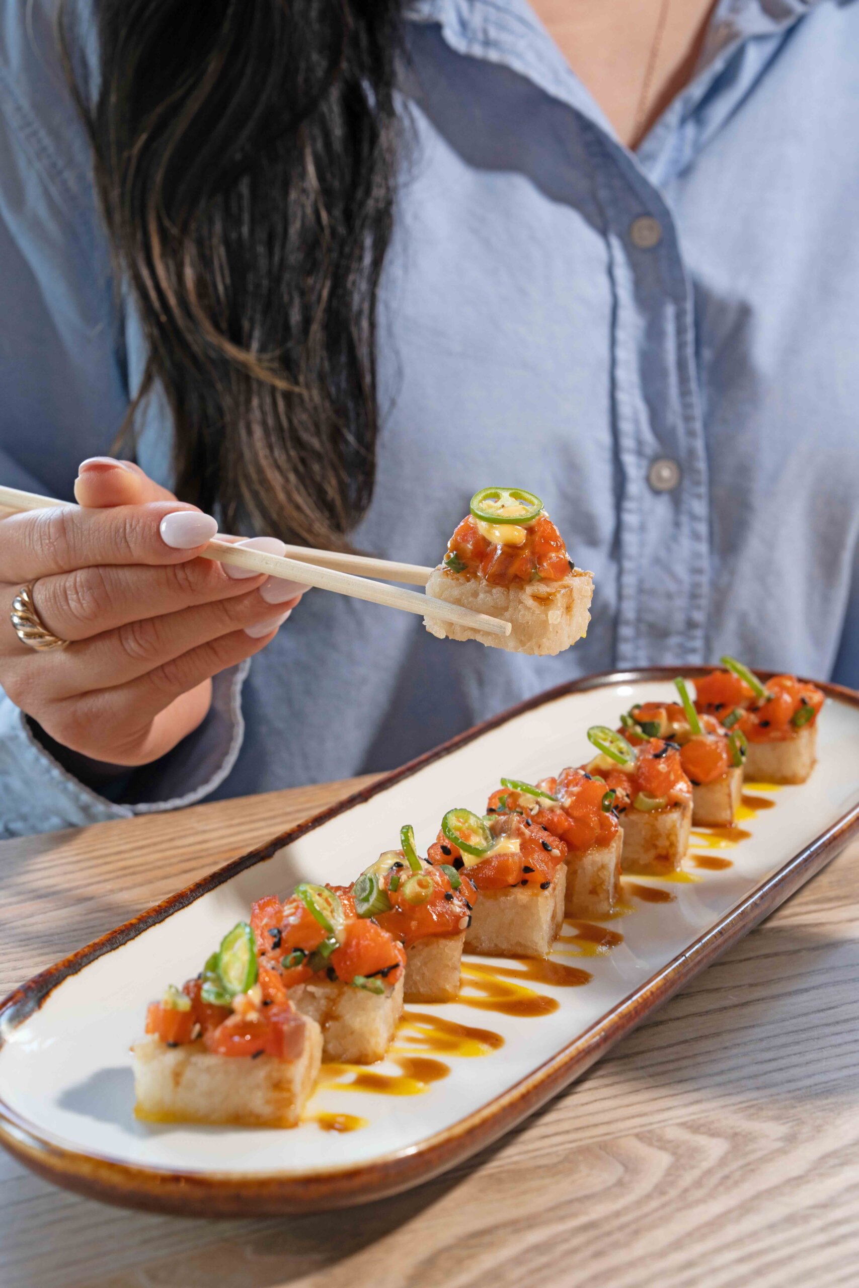 CRAFT Beer Market A person holding a piece of sushi with chopsticks above a plate of rectangular sushi pieces topped with diced fish and green garnish.