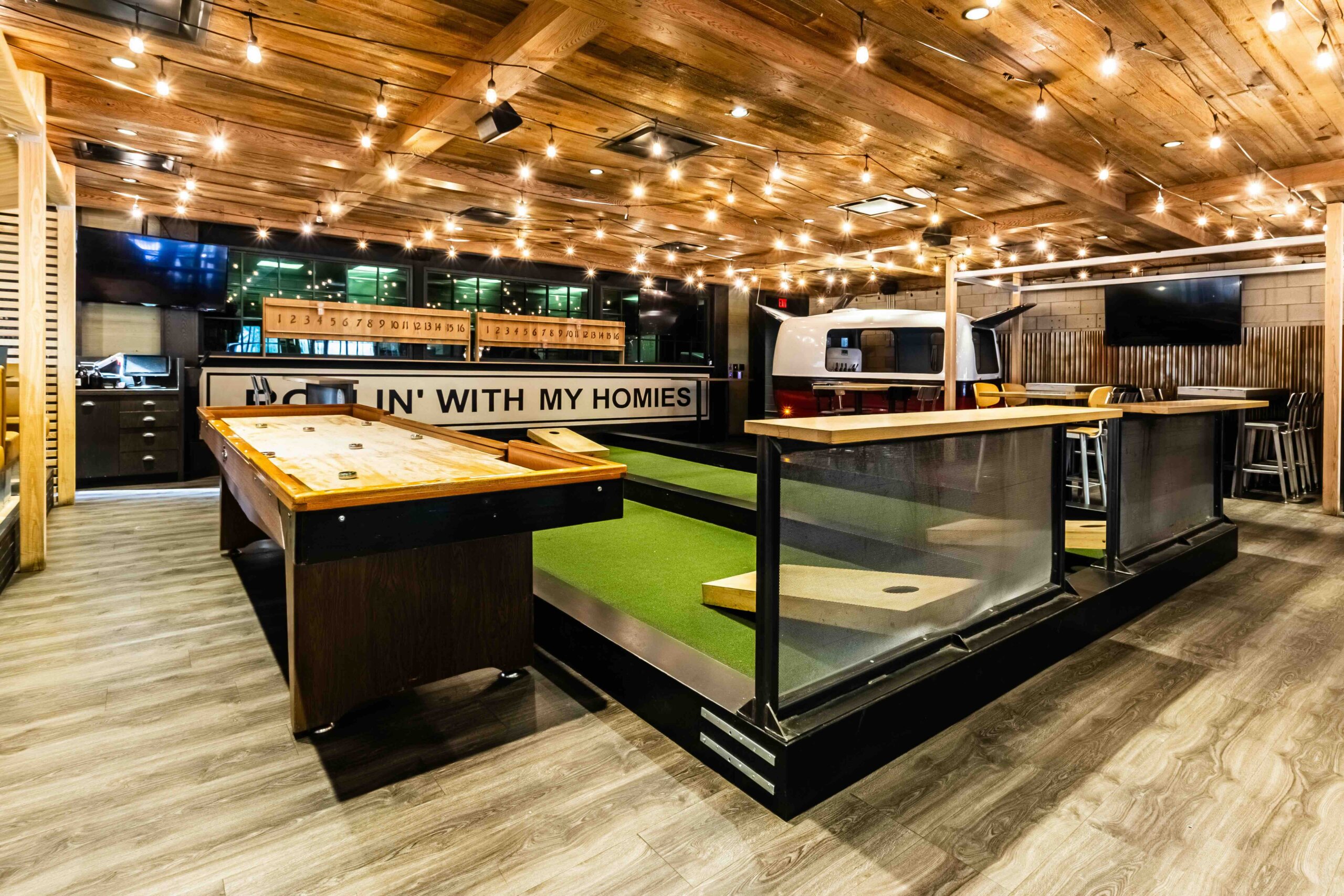 CRAFT Beer Market Indoor recreational space with string lights, a bocce ball court, shuffleboard table, seating area, and a silver trailer under a wooden ceiling.