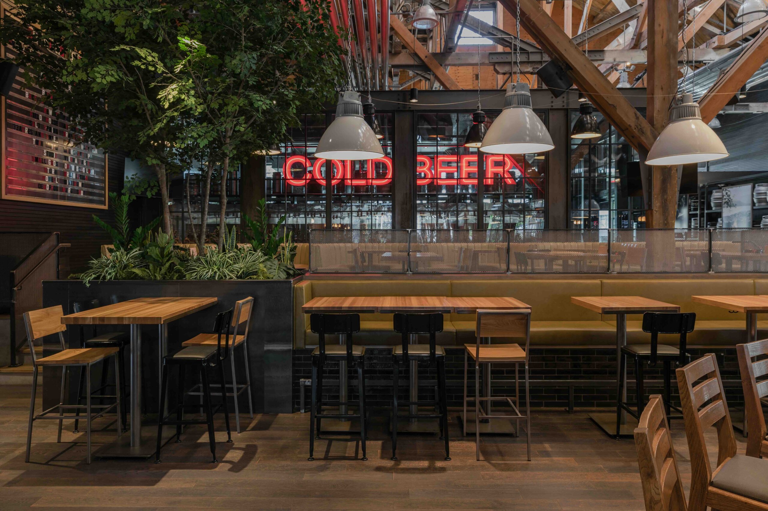 CRAFT Beer Market Spacious restaurant interior with wooden tables and chairs, indoor plants, pendant lights, and a large neon "COLD BEER" sign in the background.