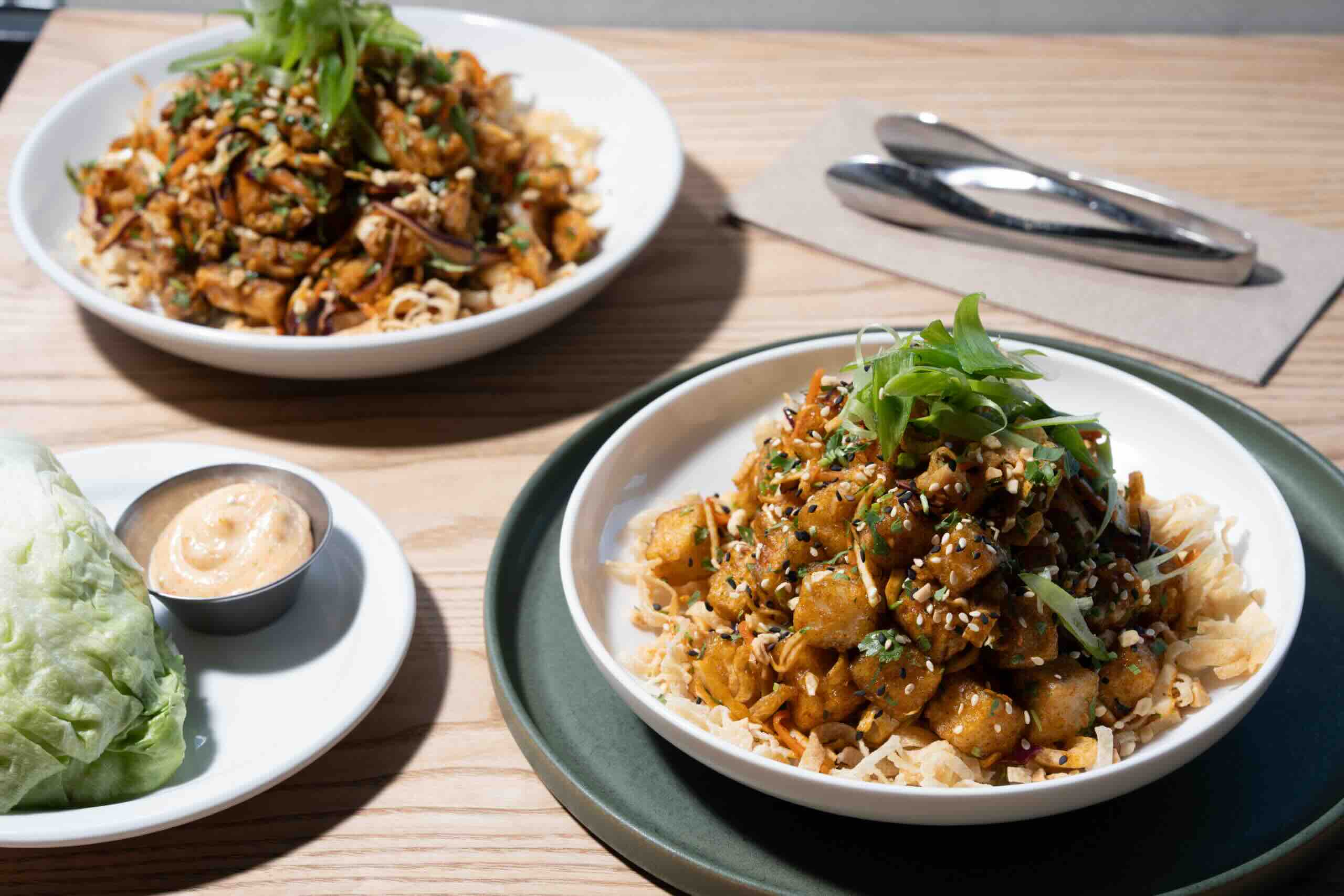 CRAFT Beer Market Two bowls of stir-fried chicken and vegetables on crispy noodles, topped with green onions and sesame seeds, served with a side of lettuce and dipping sauce—perfect to enjoy in our Beer Garden with your favorite craft beer.