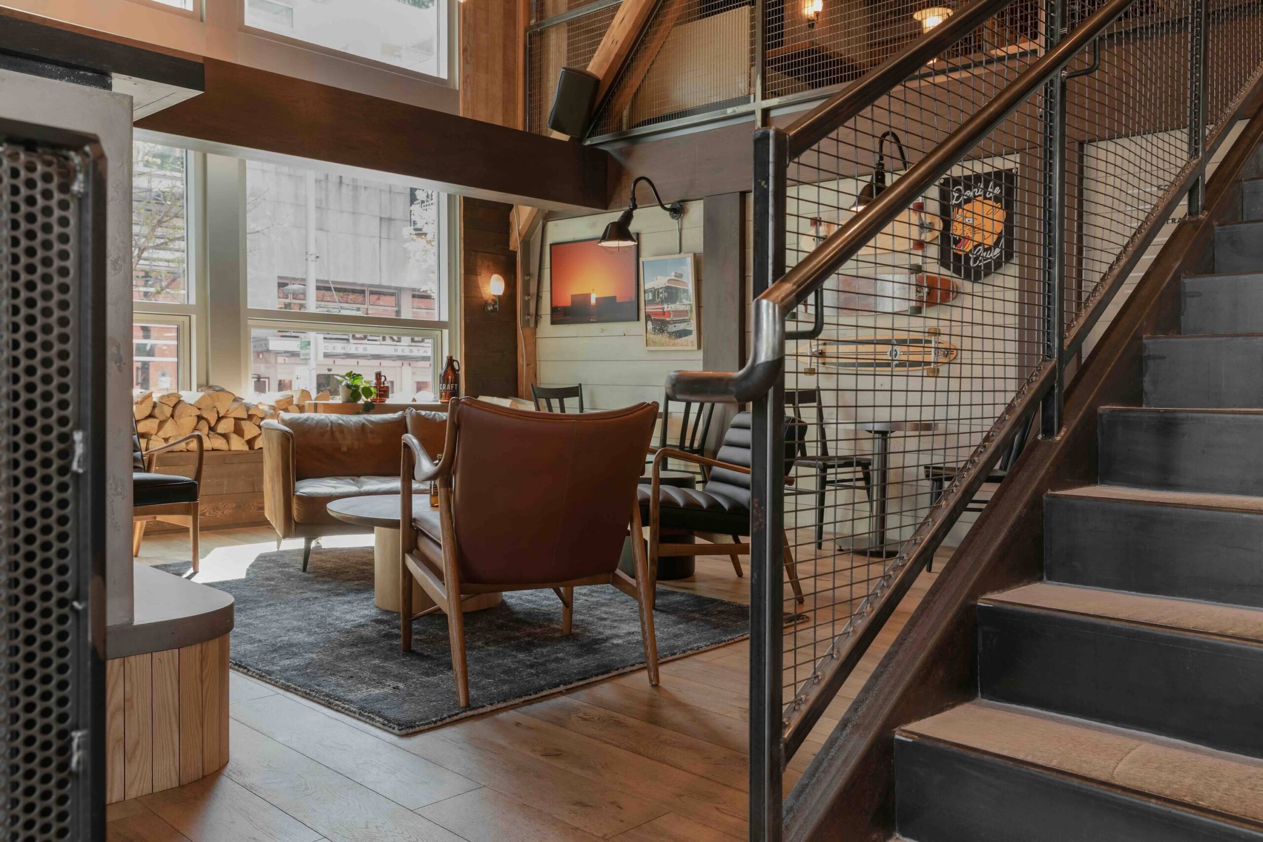 CRAFT Beer Market A modern lounge area with leather chairs, a round table, and a black rug invites social vibes next to large windows and a staircase with a metal railing.