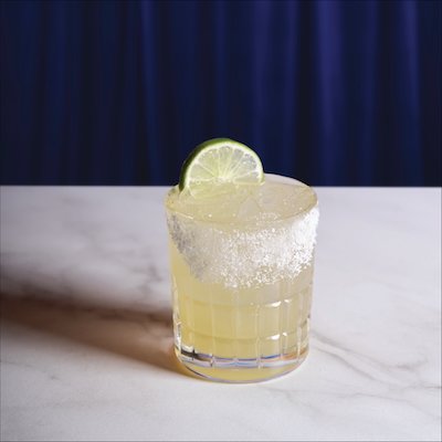 CRAFT Beer Market A margarita cocktail in a glass with a salted rim and lime wheel garnish, placed on a marble surface with a blue curtain backdrop—perfect for enjoying in a Beer Garden setting.