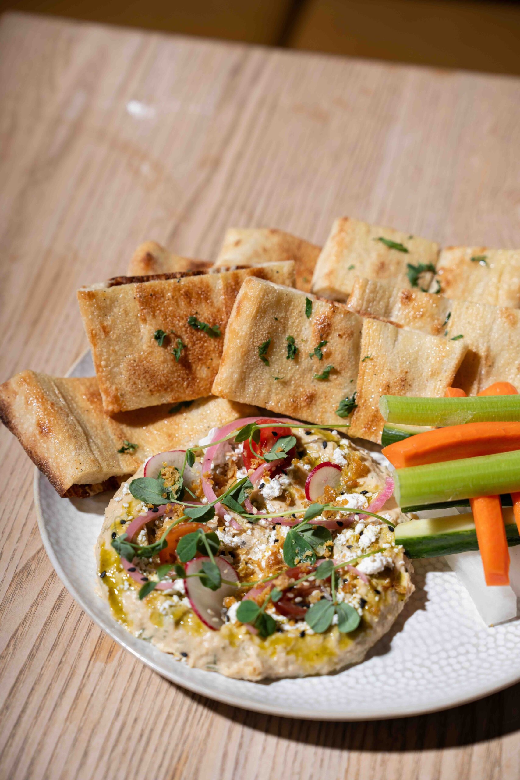 CRAFT Beer Market A plate with flatbread, hummus topped with herbs and radish slices, and vegetable sticks sits on a wooden table—perfect for sharing great food and social vibes over craft cocktails.