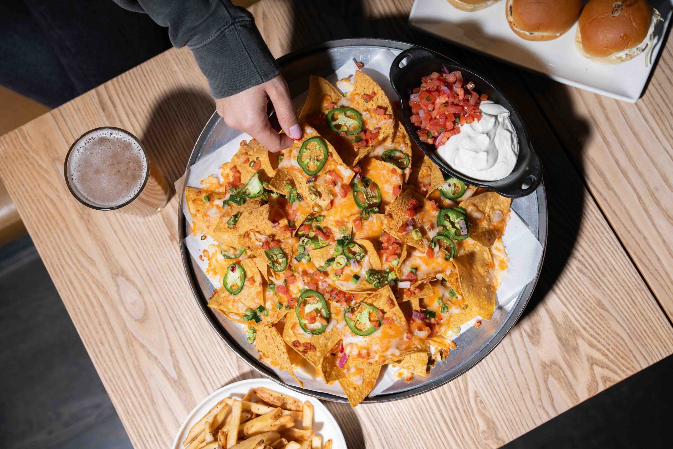 CRAFT Beer Market A person reaches for loaded nachos topped with cheese, jalapeños, and pico de gallo, served with sour cream. Fries, sliders, and a drink are also on the table.
