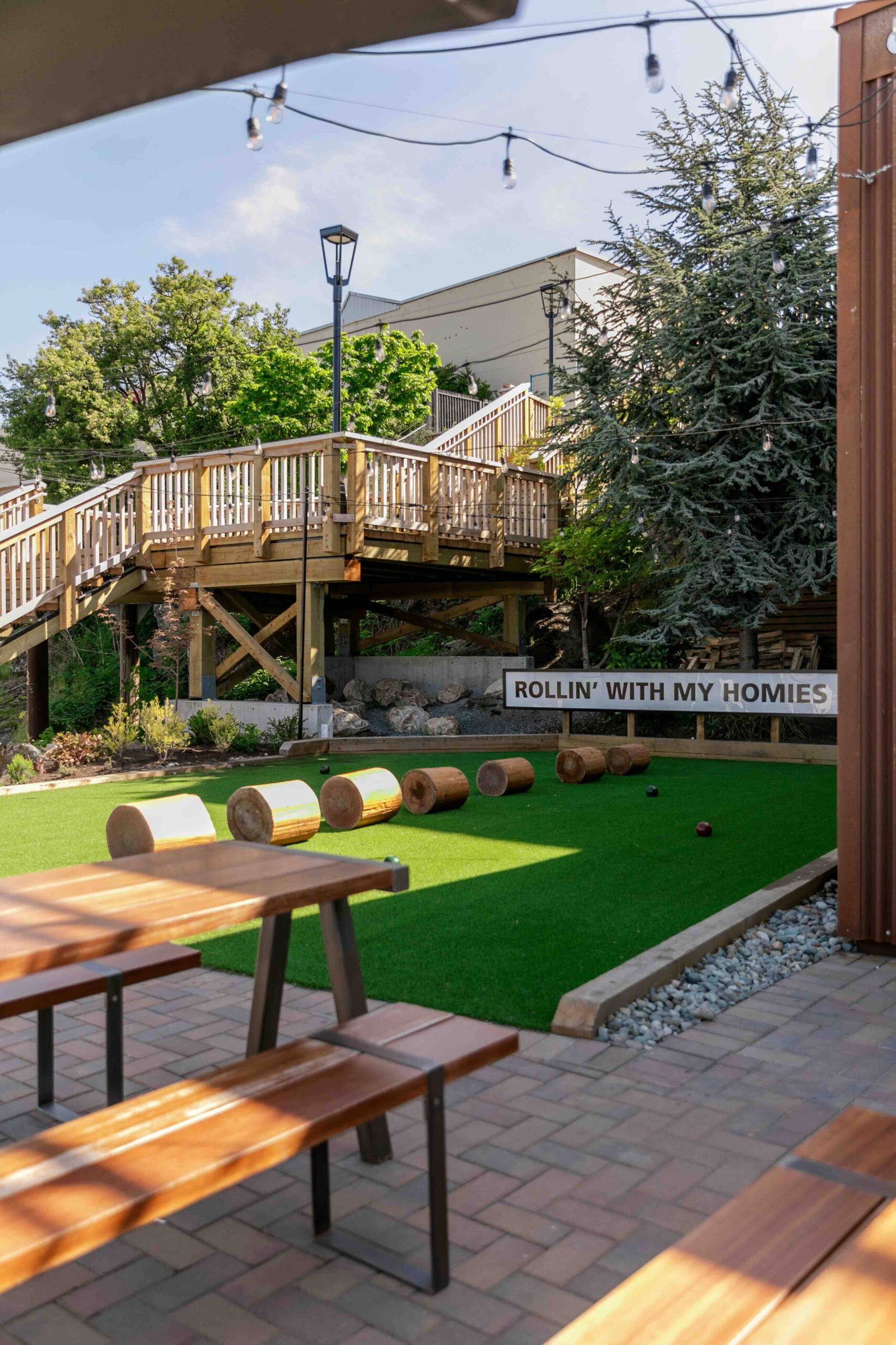 CRAFT Beer Market Outdoor seating area with picnic tables, artificial turf, log seats, string lights, and a "ROLLIN' WITH MY HOMIES" sign near a wooden walkway and trees—perfect for enjoying craft beer in this inviting beer garden.