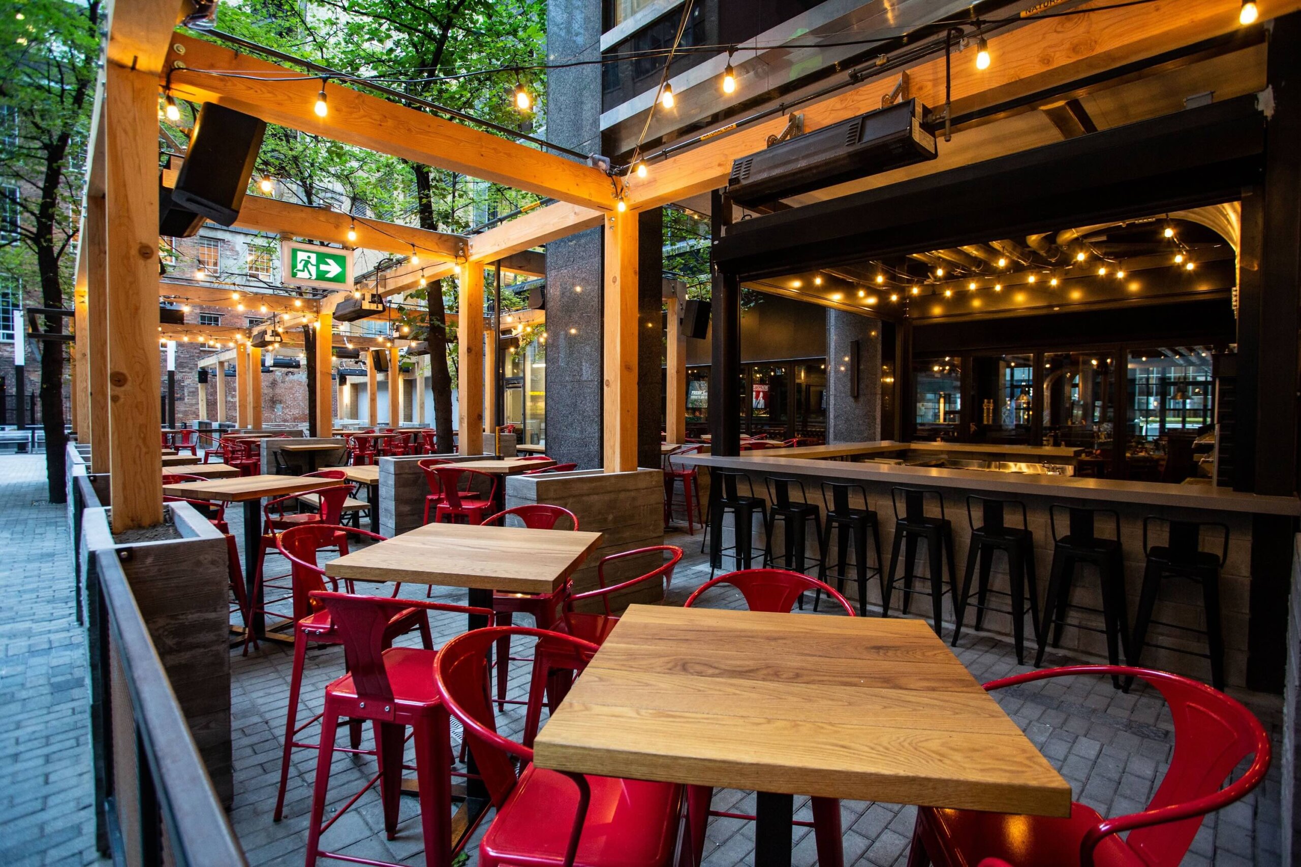 CRAFT Beer Market Outdoor patio seating area with wooden tables, red chairs, and bar stools under string lights, surrounded by trees and modern building structures.