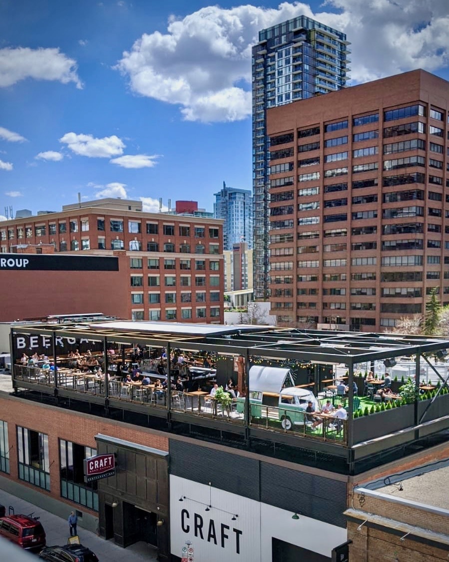 CRAFT Beer Market A rooftop restaurant with outdoor seating is busy with patrons, surrounded by tall office buildings under a partly cloudy sky in an urban area.
