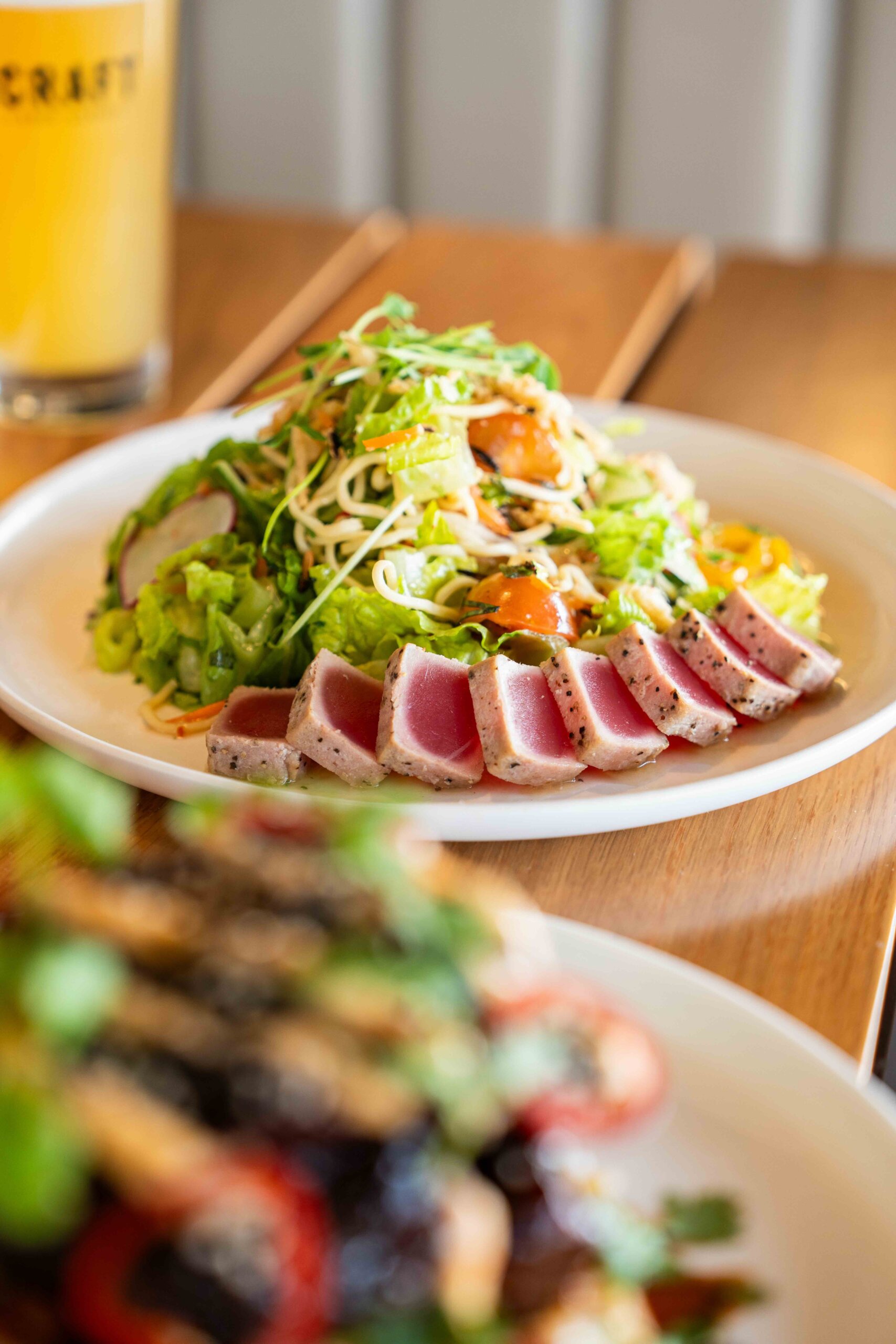 CRAFT Beer Market A plate of seared ahi tuna slices served with a mixed greens salad, cherry tomatoes, and sprouts, with a drink in the background.