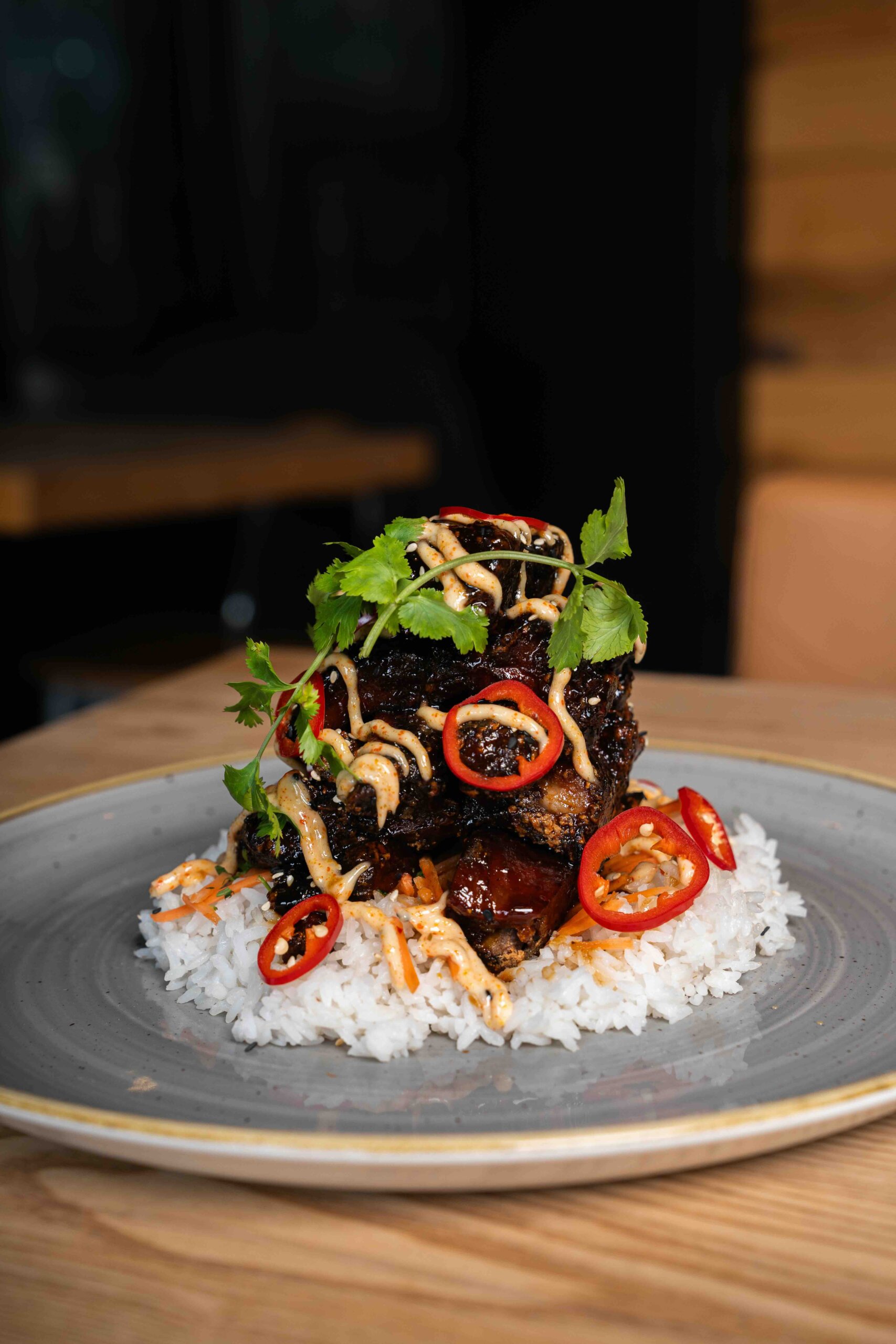 CRAFT Beer Market A serving of grilled meat topped with sauce, sliced red chili, and cilantro is placed on a bed of rice garnished with shredded vegetables—perfect for casual dining and pairing with craft cocktails in a relaxed, social vibes setting.