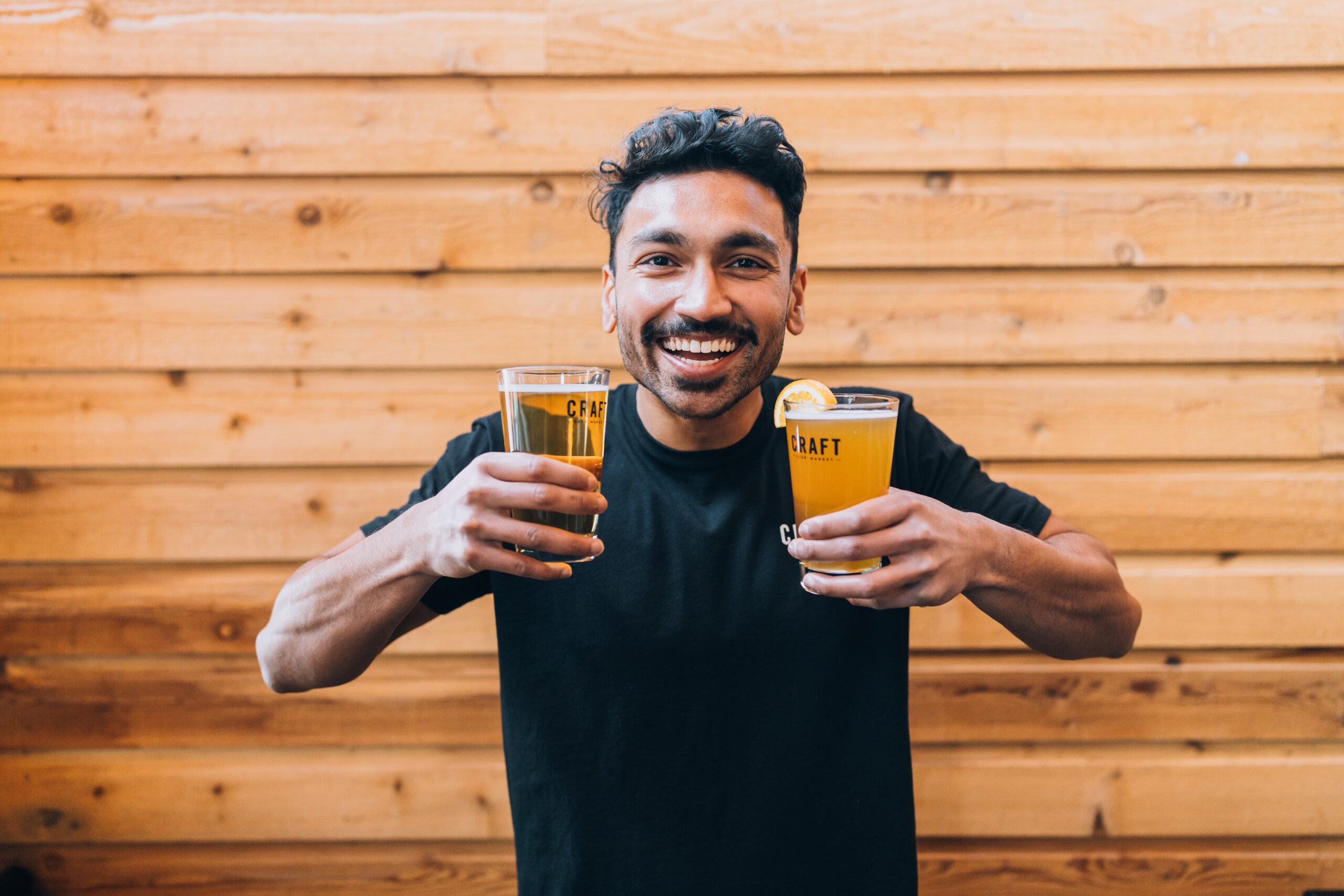 CRAFT Beer Market Smiling man in a black t-shirt holds two pints of beer in front of a wooden wall.