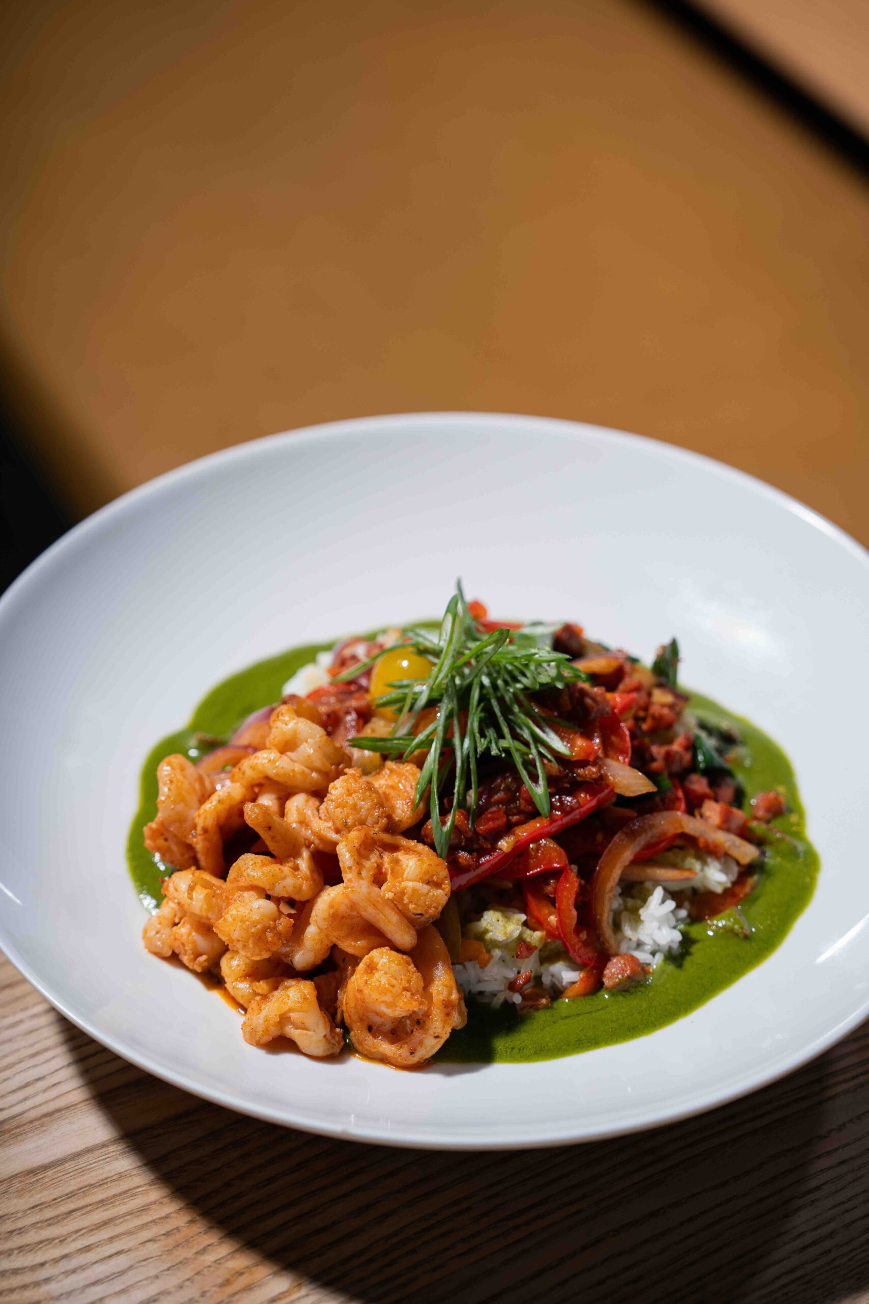 CRAFT Beer Market A white bowl contains roasted cauliflower, rice, sautéed vegetables, a green sauce, and fresh herbs, perfect for enjoying in a casual dining setting on a wooden table.