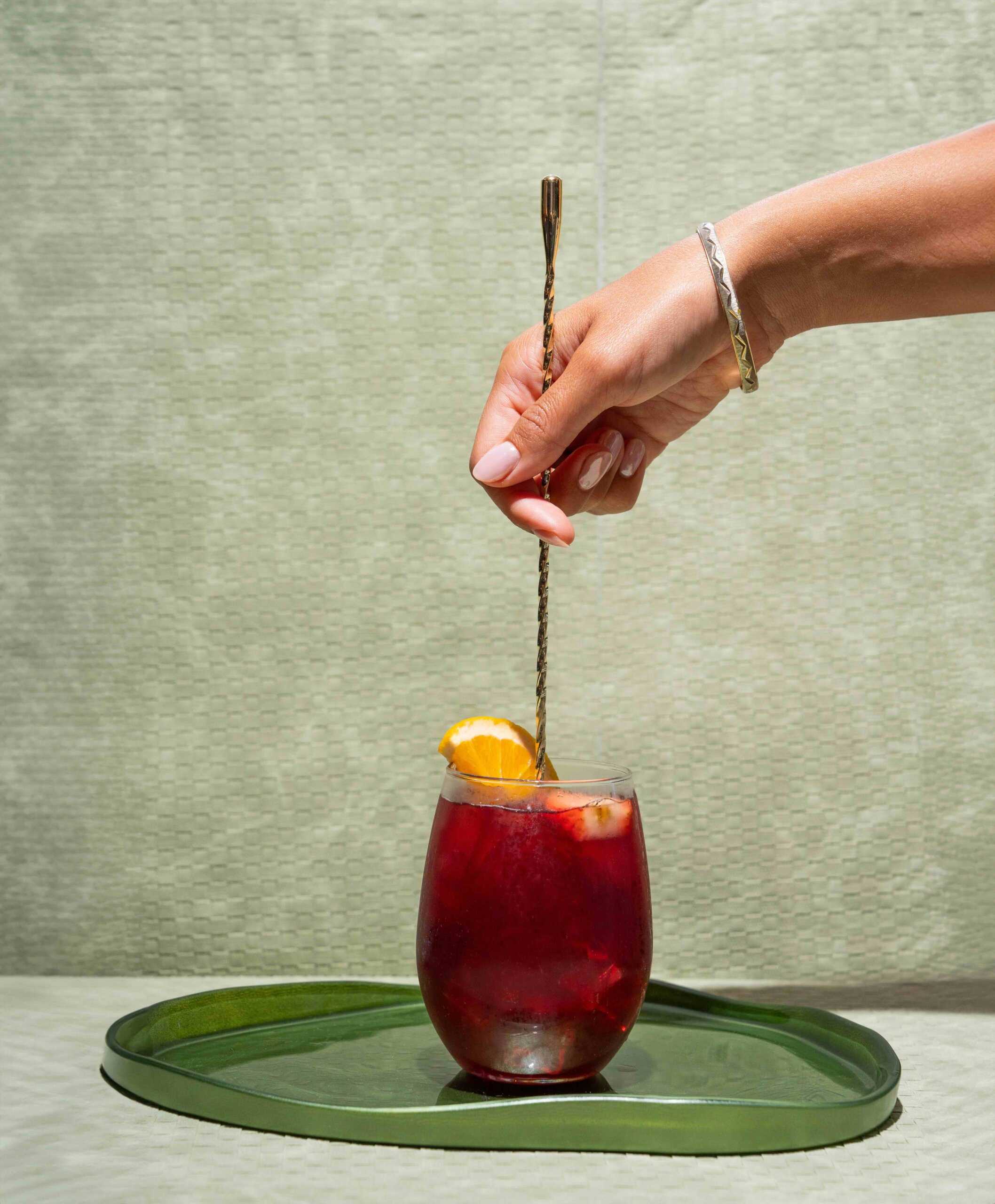 CRAFT Beer Market A hand stirs a glass of red drink with a spiral spoon; orange slices are used as garnish. The glass sits on a green tray against a light textured background.