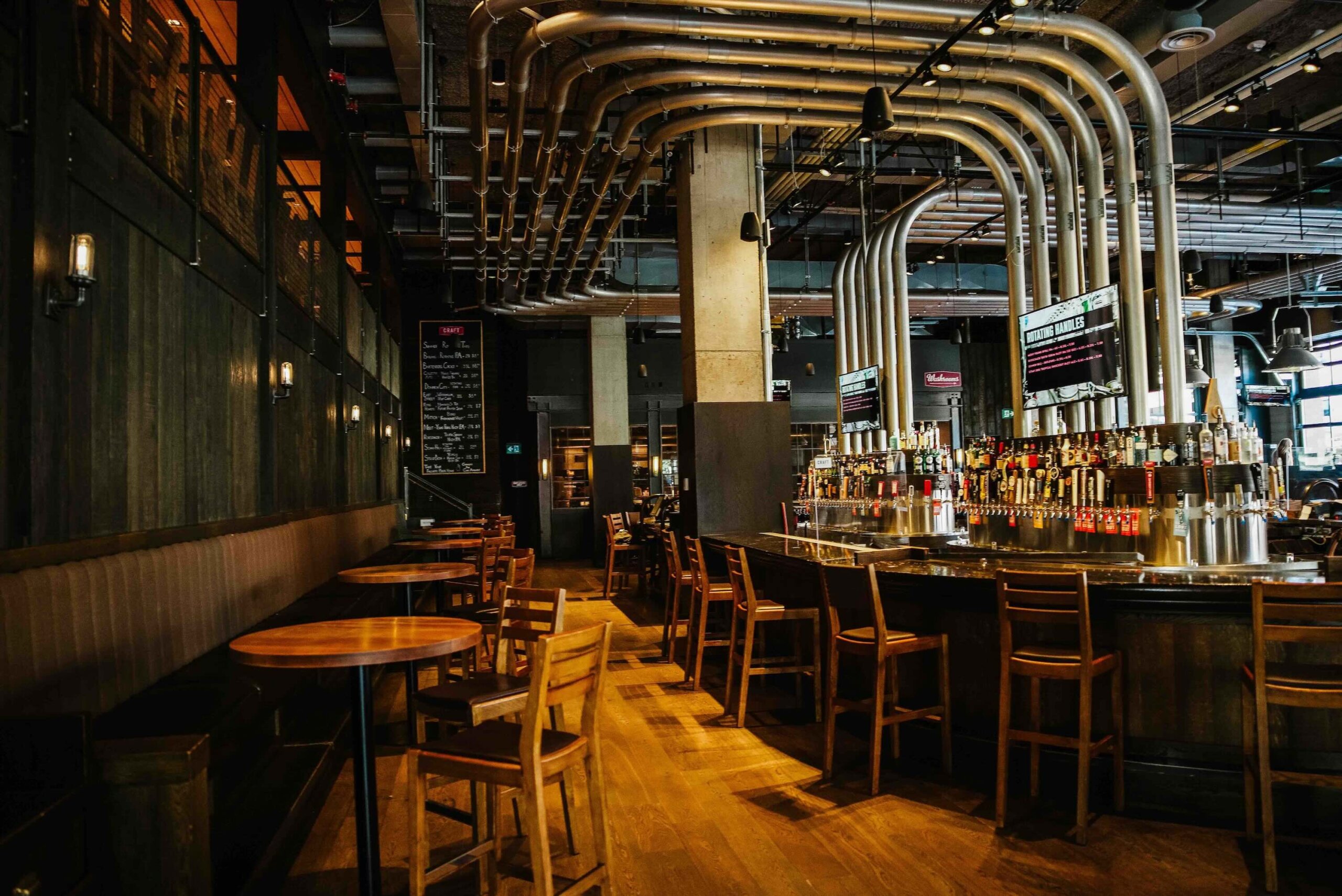 CRAFT Beer Market A modern bar with wooden tables and chairs, a long bar counter, and multiple metallic pipes overhead in an industrial-style interior.