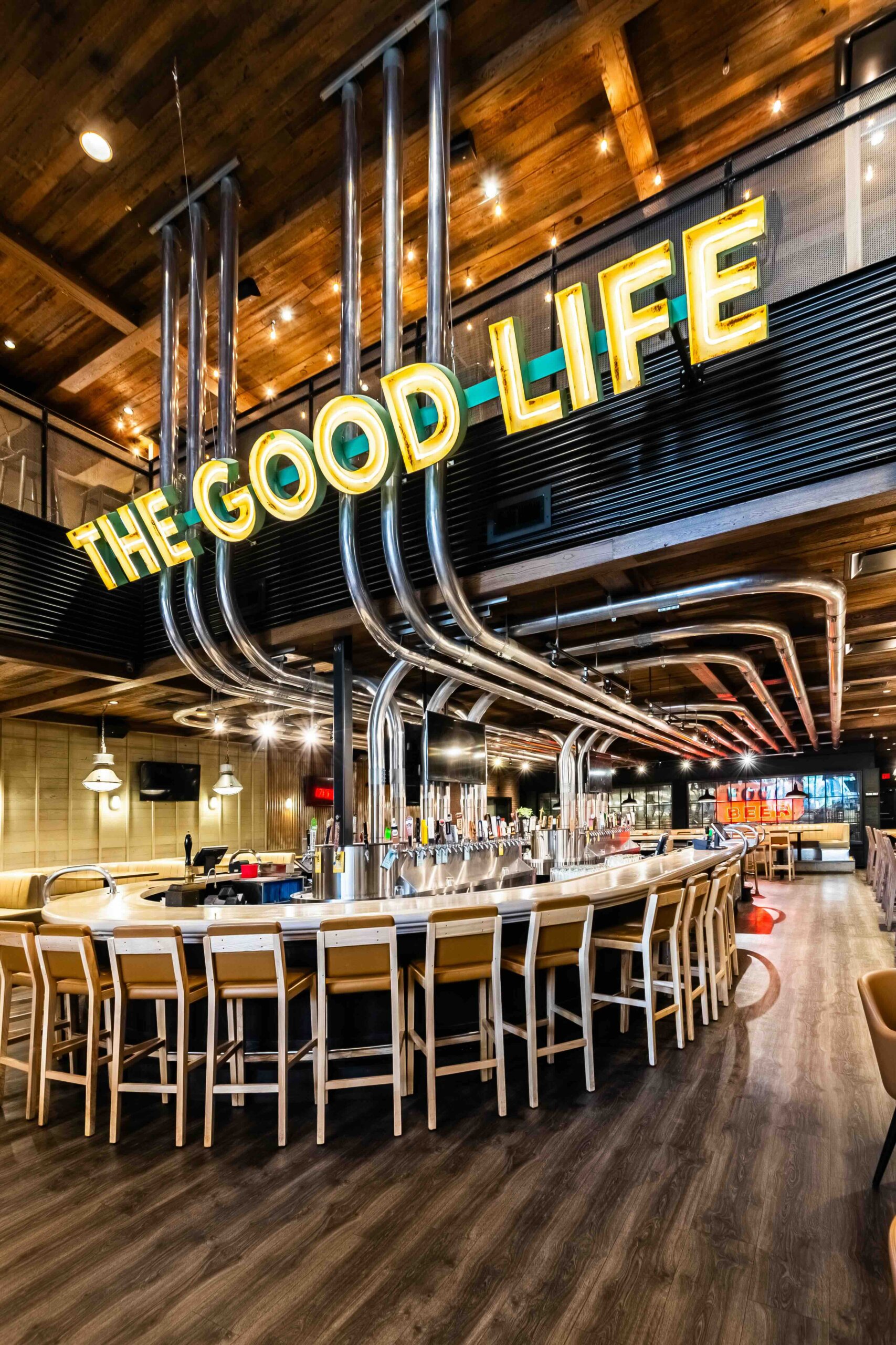 CRAFT Beer Market A modern bar with wooden floors, high chairs around a curved counter, and a large neon sign reading “THE GOOD LIFE” above overhead metal pipes.