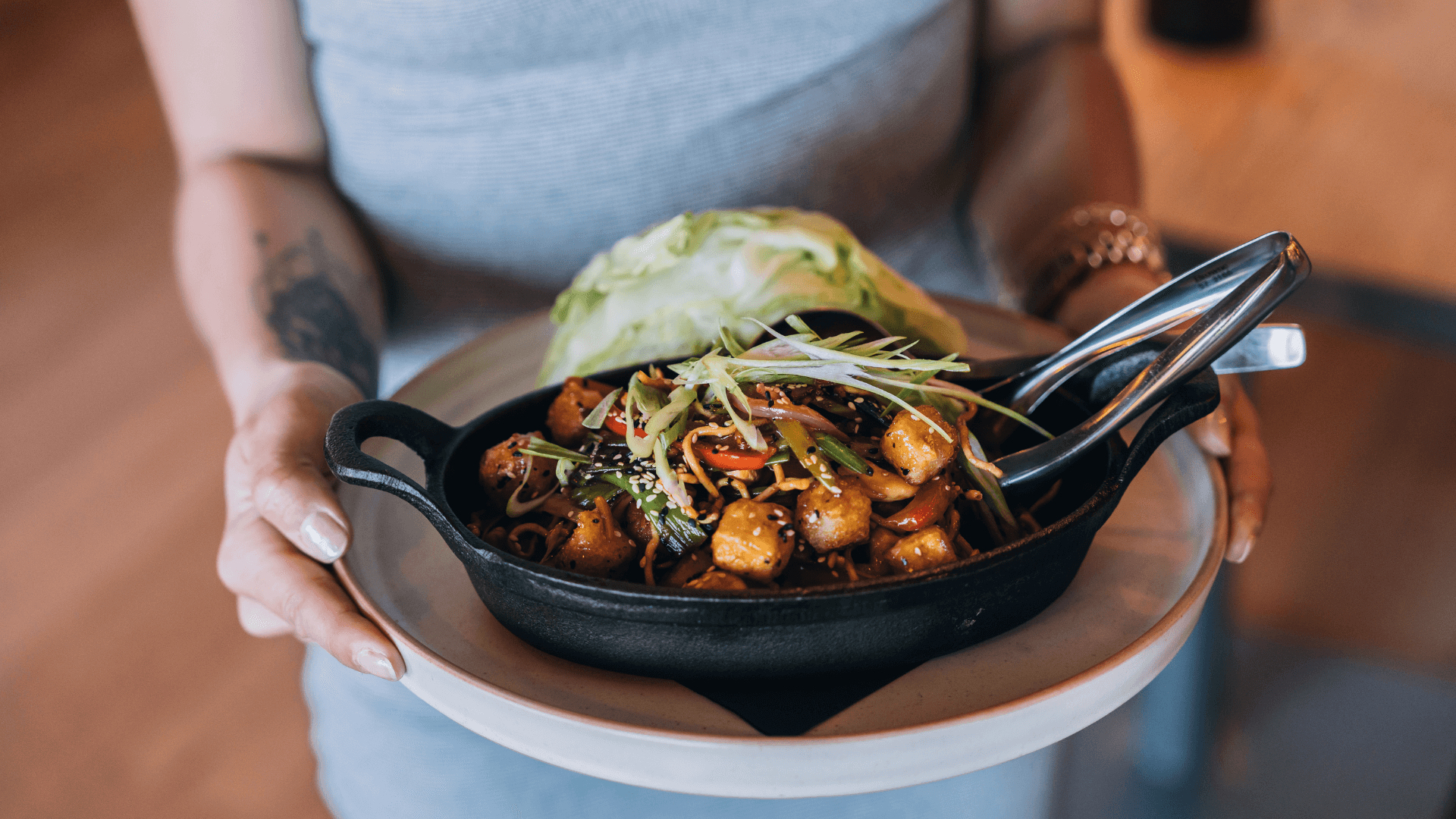 CRAFT Beer Market A person holds a plate with a cast iron skillet containing sautéed tofu, vegetables, and sliced green onions next to a wedge of lettuce—perfect to enjoy alongside our signature craft cocktails.