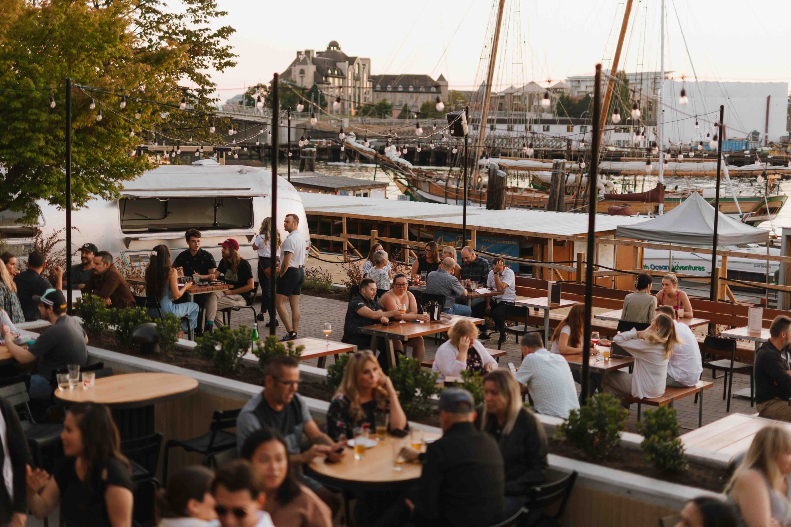 CRAFT Beer Market People sit at outdoor tables by a waterfront beer garden, enjoying casual dining and social vibes as boats are docked in the background on a sunny day.