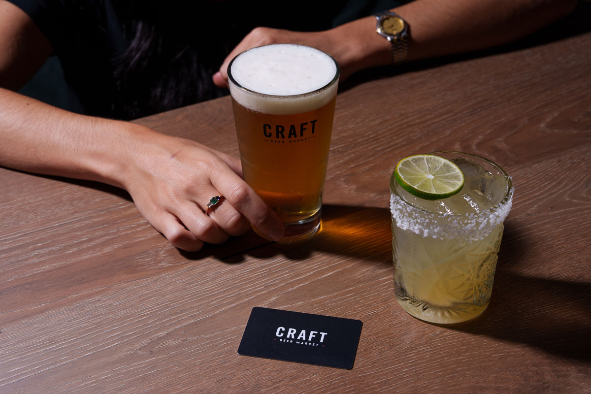 CRAFT Beer Market A person holds a pint of craft beer labeled "Craft" next to a salt-rimmed craft cocktail with a lime slice, on a wooden table with a "Craft" card.
