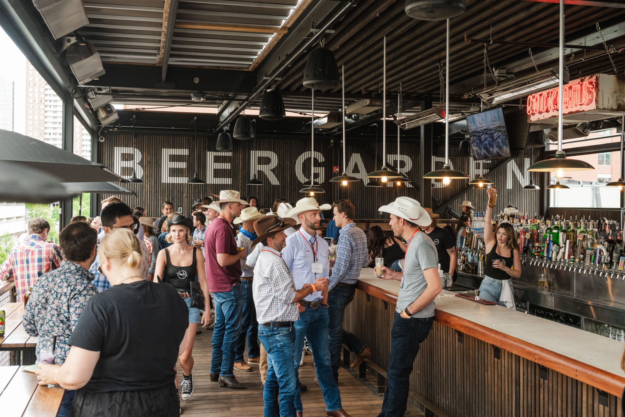 CRAFT Beer Market A group of people wearing casual and western-style clothing gather and converse at an open-air bar labeled “BEER GARDEN,” enjoying craft beer under the night sky.