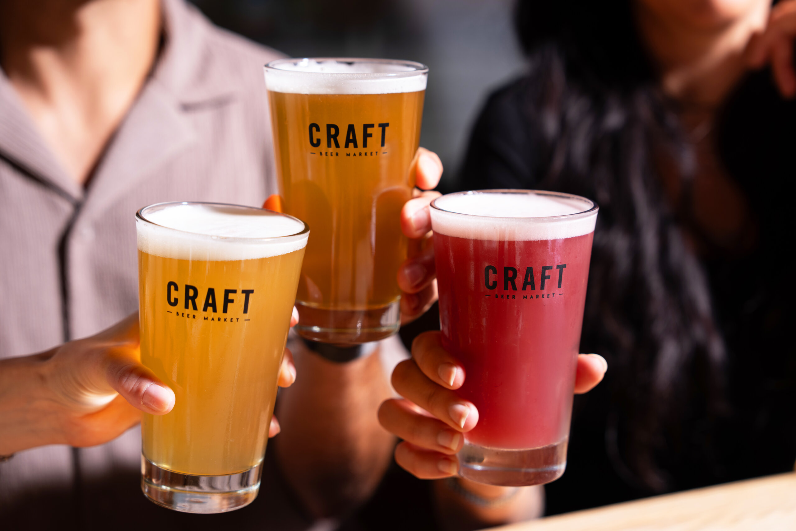 CRAFT Beer Market Three people hold up glasses of beer, two light and one reddish, all labeled "Craft Beer Market," in a cheers gesture, perfect for group bookings or private events.
