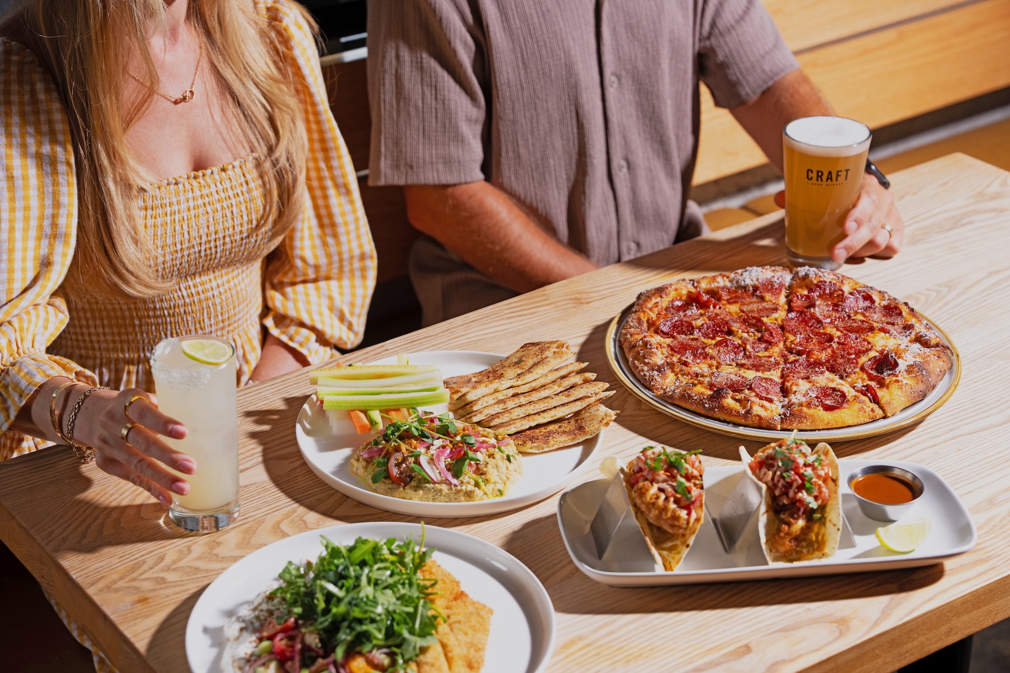 CRAFT Beer Market Two people sit at a wooden table enjoying pizza, tacos, dip with celery and flatbread, salad, lemonade, and a pint of beer—all made with locally sourced ingredients.