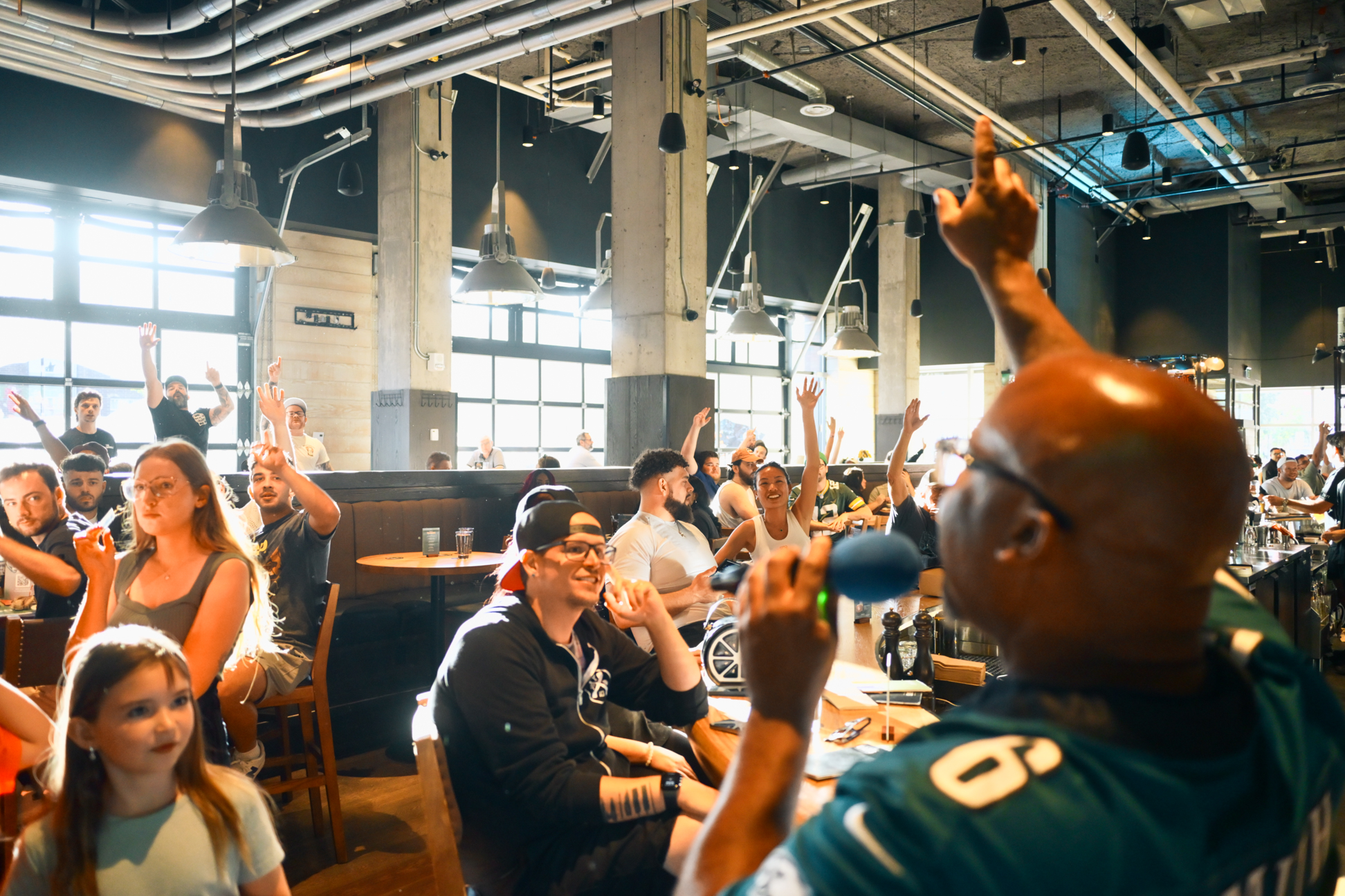CRAFT Beer Market A man with a microphone engages an enthusiastic crowd at a busy indoor event, where several people have their hands raised—perfect for private events featuring craft beer and lively entertainment.