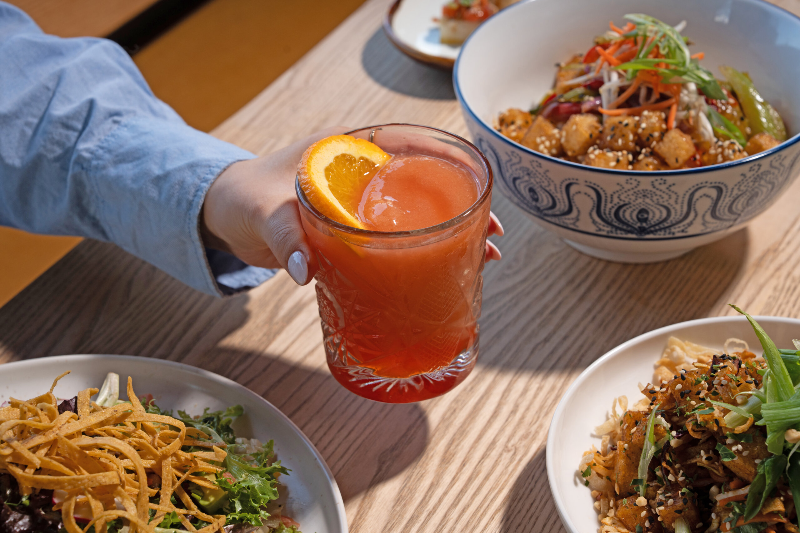 CRAFT Beer Market A person holding a glass of craft cocktail with an orange slice, surrounded by plates of salad and Asian-inspired dishes on a wooden table.