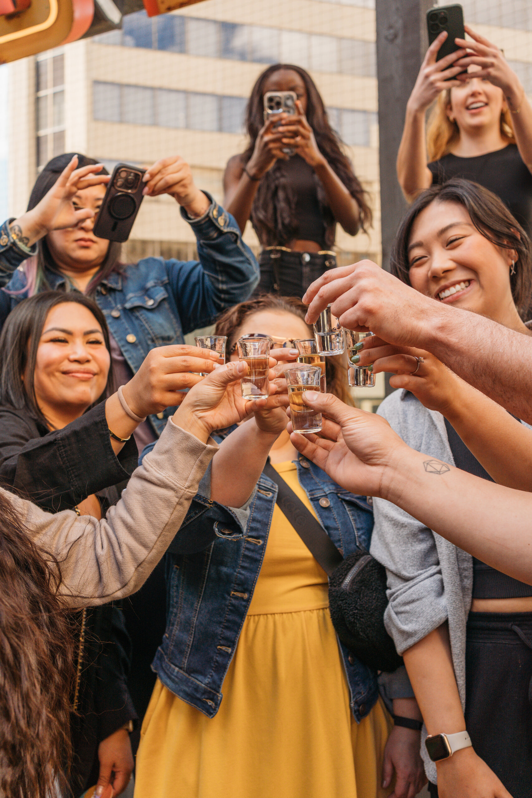 CRAFT Beer Market A group of people standing close together outdoors, raising shot glasses in a toast with craft cocktails, while some take photos with their phones.