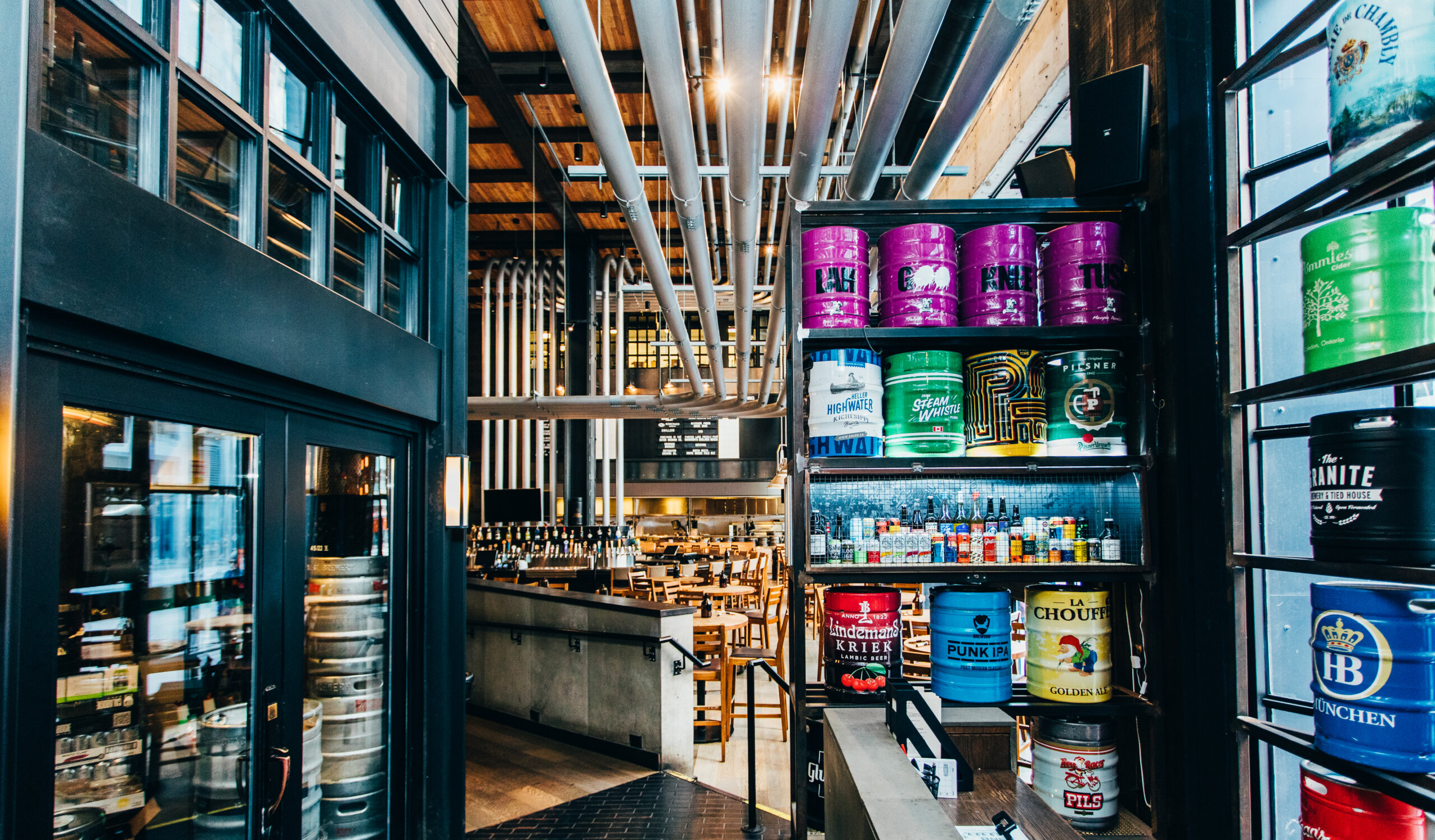 CRAFT Beer Market Interior of a modern brewery with colorful beer kegs on shelves, large metal pipes overhead, and tables set up in the background—perfect for group bookings and gatherings.