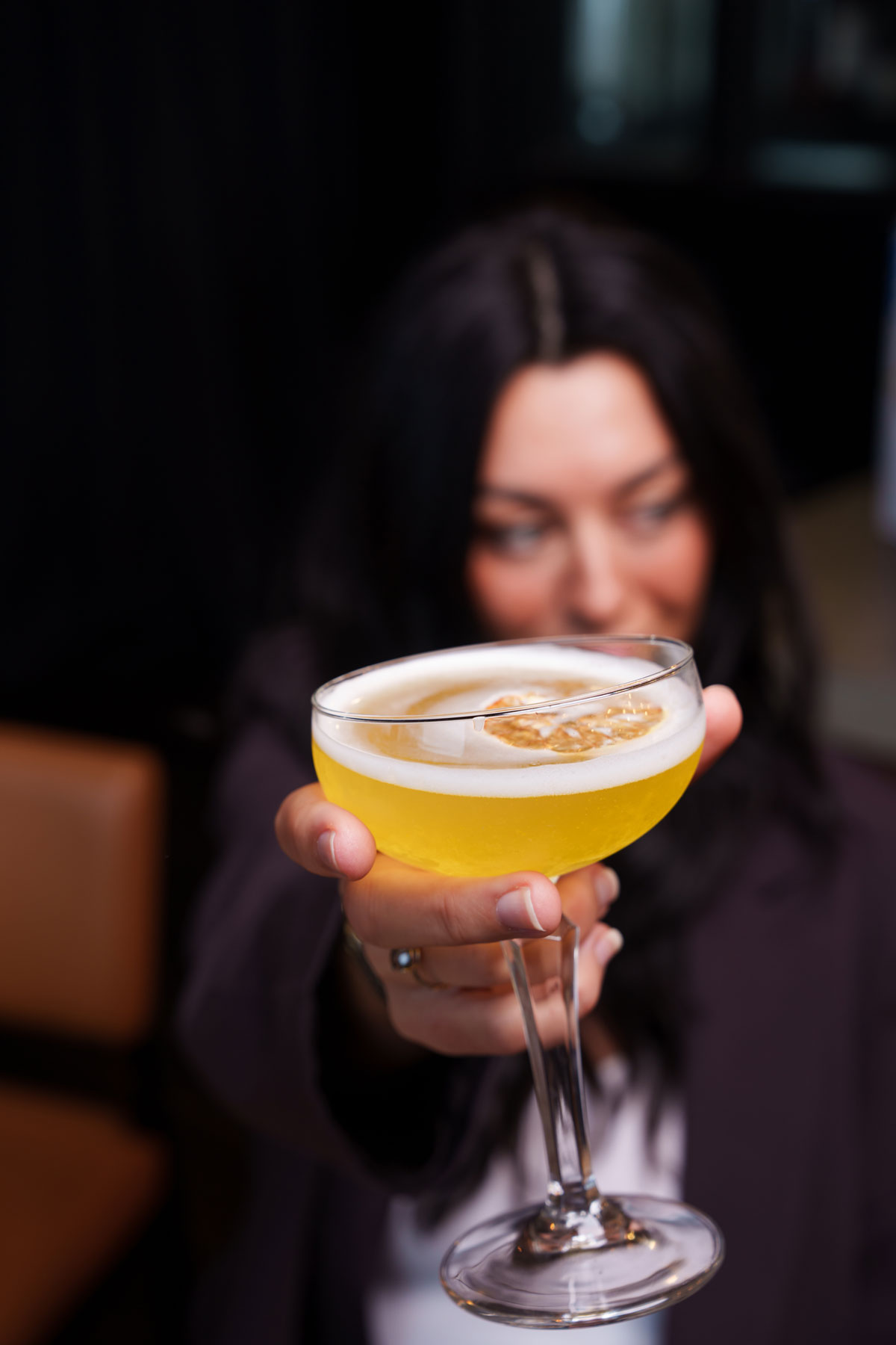 CRAFT Beer Market A person with long dark hair holds a coupe glass filled with a yellow cocktail topped with foam and a dried garnish, focusing the drink toward the camera—showcasing a sustainable twist on craft beer-inspired cocktails.