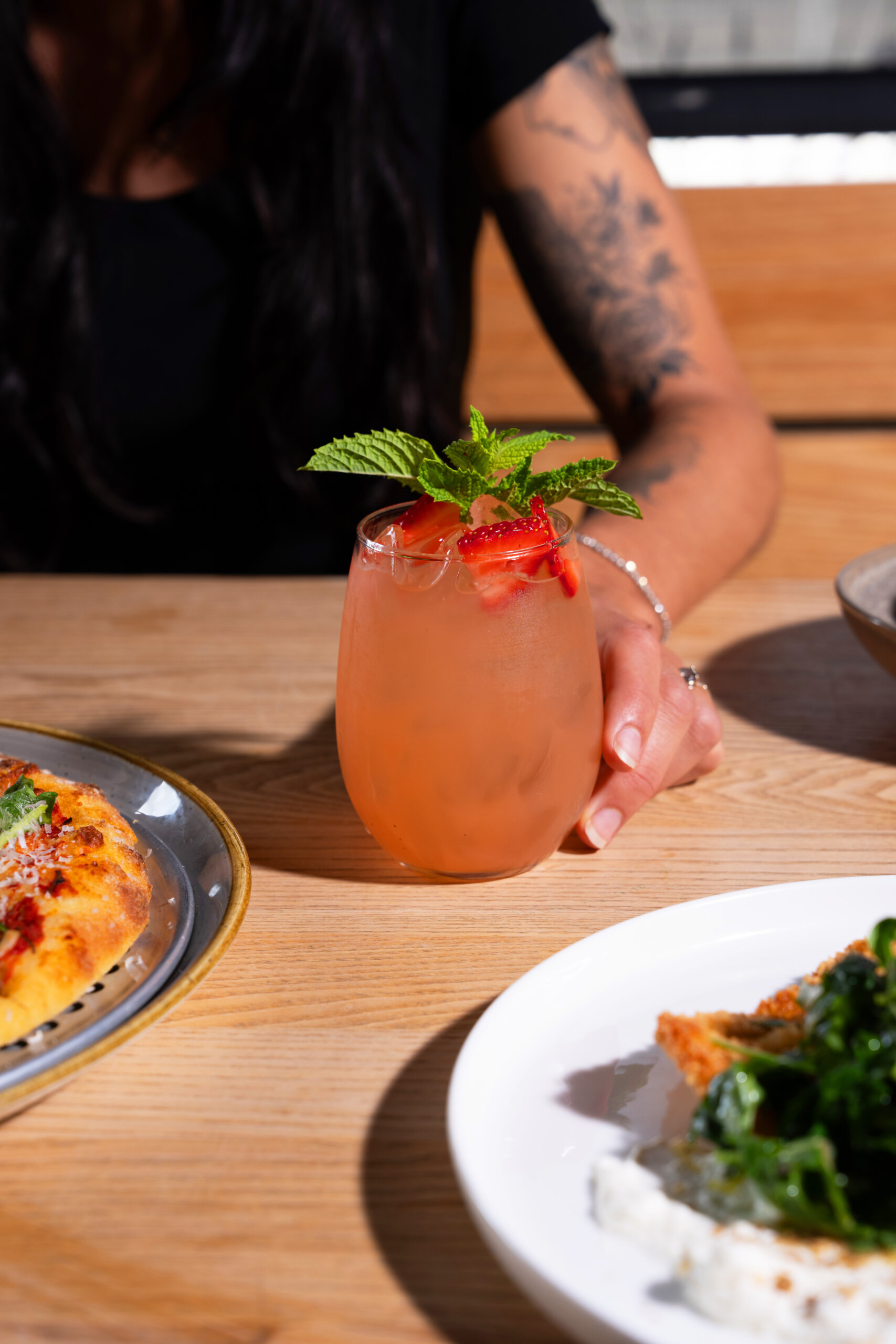 CRAFT Beer Market A person with a tattooed arm holds a glass of pink craft cocktail garnished with mint and strawberries, next to plates of pizza and salad on a wooden table, setting the scene for laid-back social vibes.